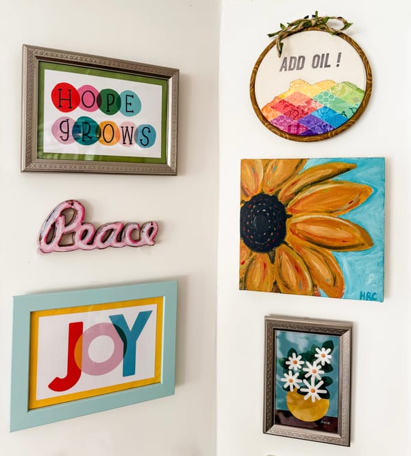 Six pieces of colorful art on a white wall. Text on the art: Hope Grows, Add Oil, Peace, Joy, and Here. 